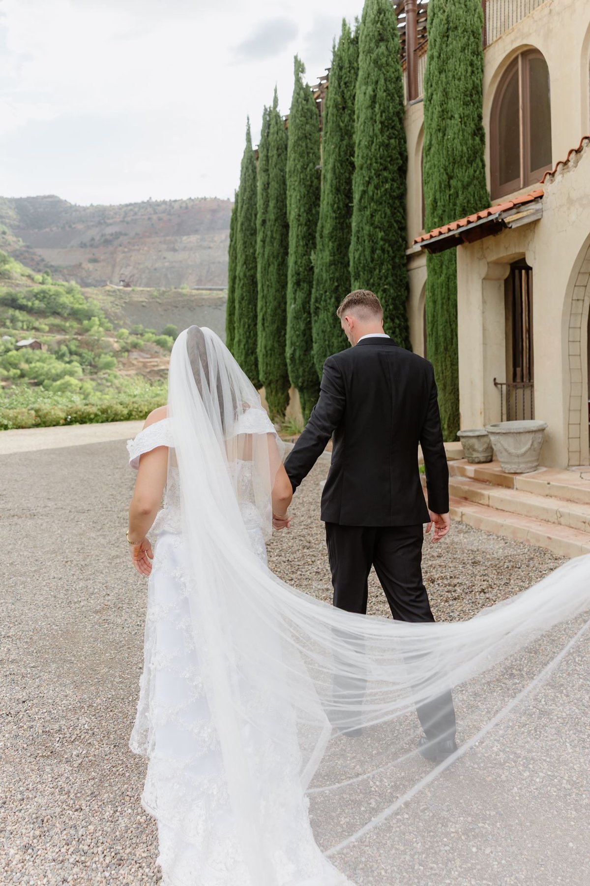 Abigail and Ethan walking beside cypress trees with her veil trailing behind