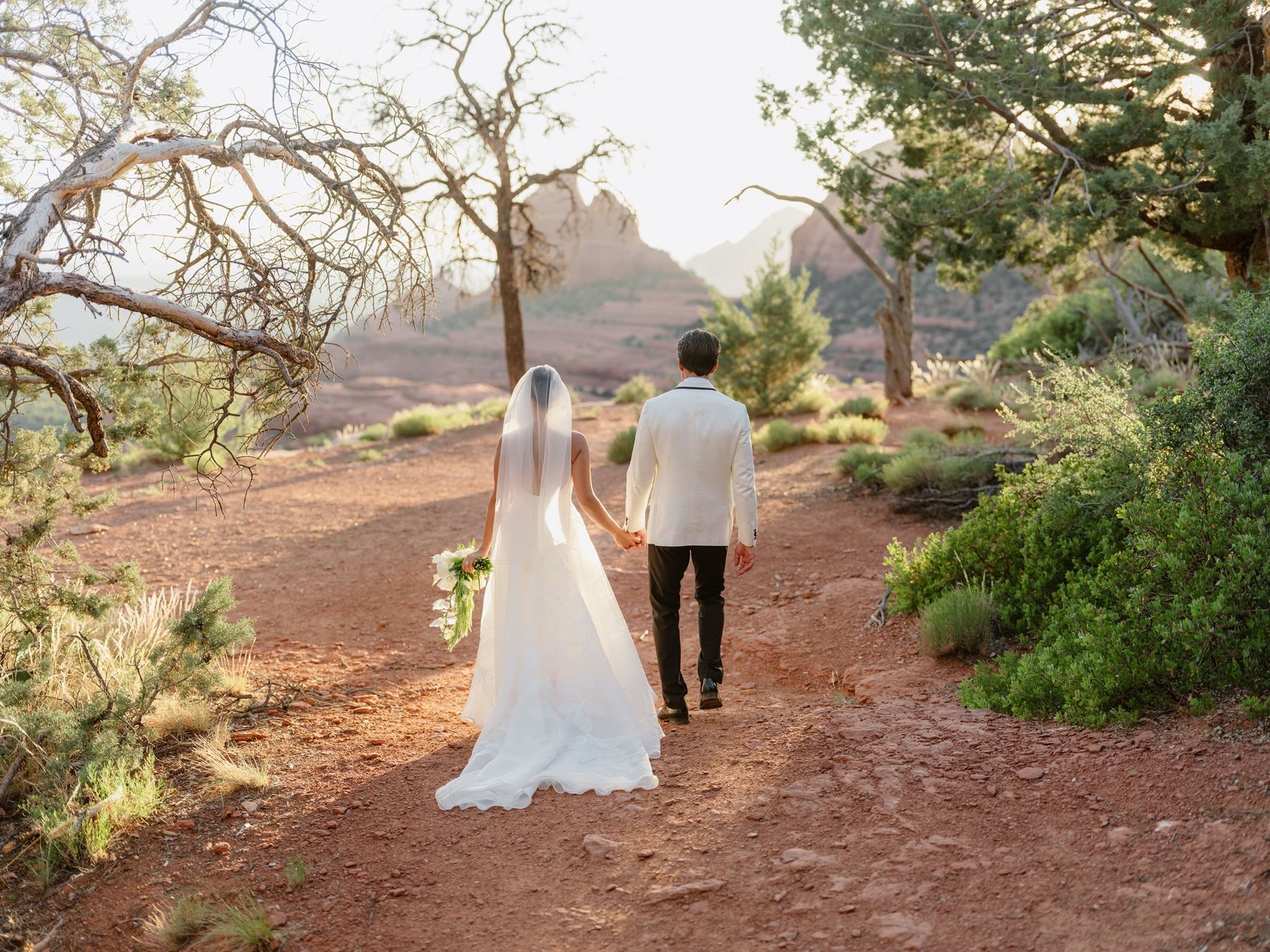 Maggie and Brandon walking hand in hand along a red dirt path beneath Sedona trees