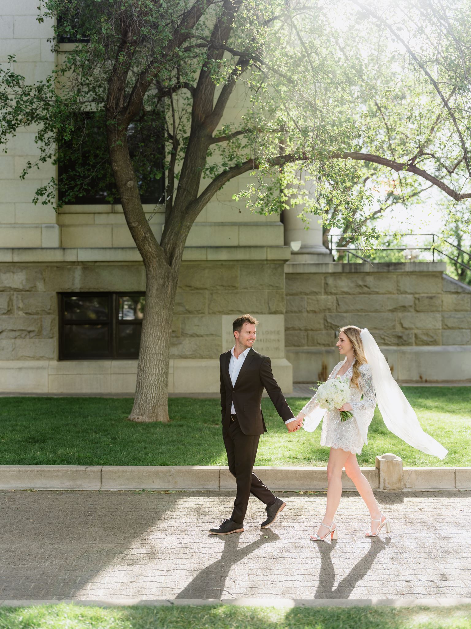 Erica and Evan walking past the courthouse with her veil trailing behind.